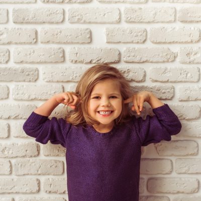 Beautiful little girl in casual clothes is covering her ears, looking at camera and smiling, standing against white brick wall
